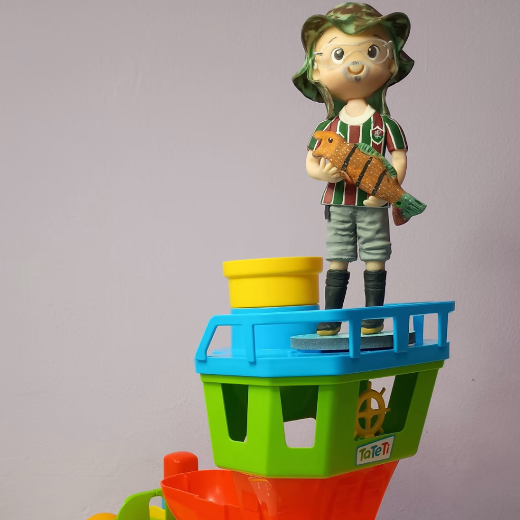 Boneco de pescador de biscuit em cima de barquinhos coloridos empilhados, com balde amarelo