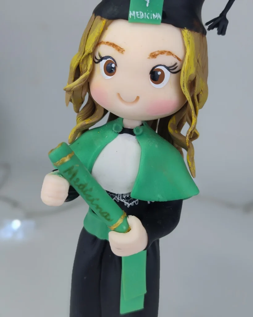 Boneca de biscuit formanda com capelo, beca preta e faixa verde, segurando o canudo