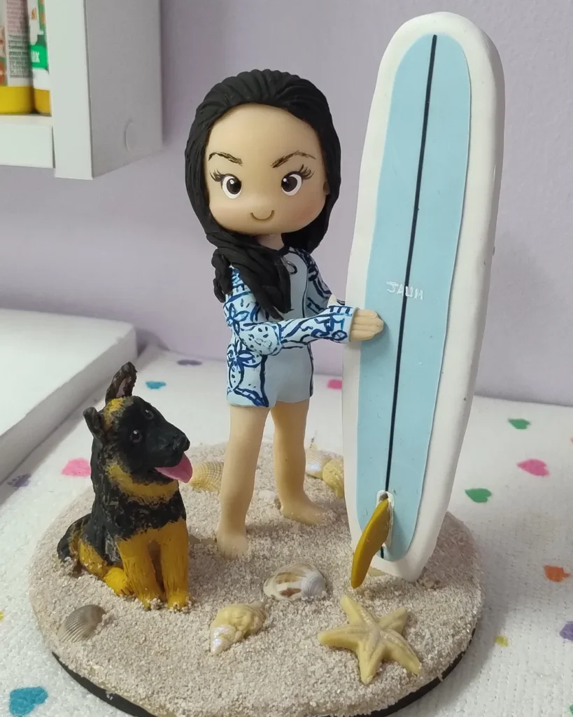 Miniatura de biscuit de menina com prancha de surf e cachorro pastor alemão na areia