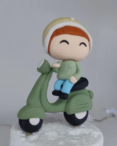 Miniatura de biscuit de bebê em scooter verde, com touca e olhos fechados, sorridente