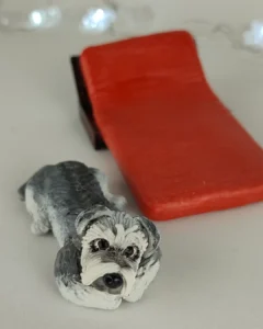 Cachorrinho de biscuit, cinza e branco, deitado no chão ao lado de uma espreguiçadeira vermelha em miniatura.