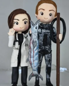 Topo de bolo de casal em biscuit: ela médica de jaleco e estetoscópio, ele pescador de roupa de mergulho com um peixe grande