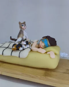Cena em biscuit de pessoa deitada na cama com máscara e dois gatos ao lado