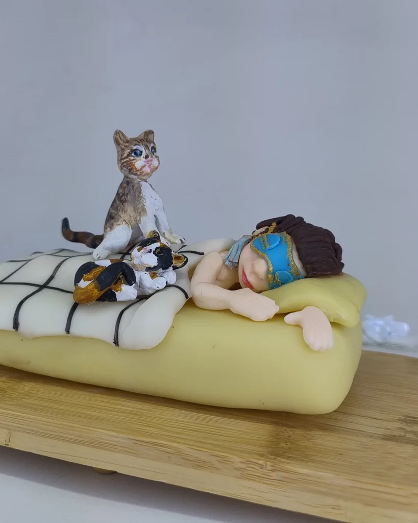 Cena em biscuit de pessoa deitada na cama com máscara e dois gatos ao lado