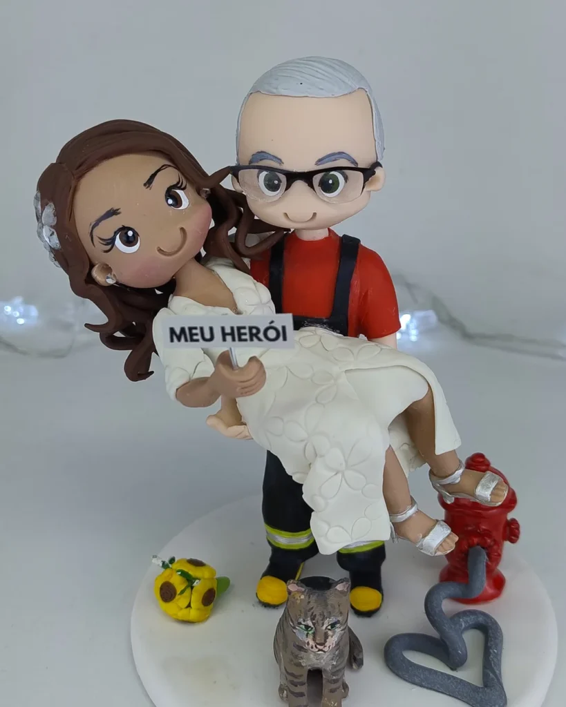 Topo de bolo de casamento em biscuit com a noiva no colo do noivo bombeiro, placa "Meu herói", hidrante e o gatinho do casal