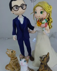 Topo de bolo de casamento em biscuit com noivo de óculos, noiva loira com buquê e três gatinhos na base