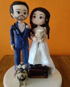 Topo de bolo de casamento em biscuit personalizado: noivo médico de terno azul, noiva com buquê sobre livros e o cachorrinho do casal